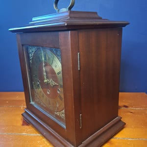 May include: A rectangular, dark brown wooden clock with a brass handle and a gold-colored clock face. The clock face has Roman numerals and decorative detailing. The clock is set on a wooden surface.