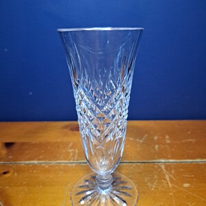 Puede incluir: Una copa de champ&aacute;n transparente, tallada, con un patr&oacute;n de diamantes y una base redonda. El vaso tiene un tallo largo y una parte superior acampanada. El vaso est&aacute; sobre una superficie de madera con un fondo azul.