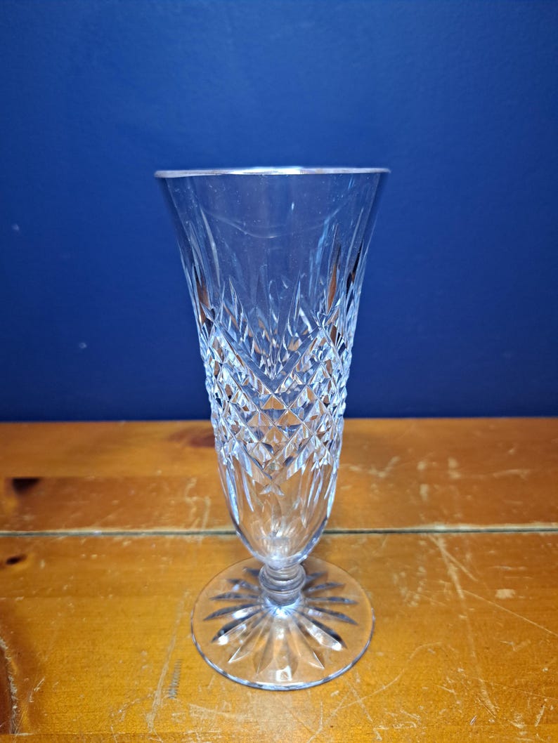 Puede incluir: Una copa de champ&aacute;n transparente, tallada, con un patr&oacute;n de diamantes y una base redonda. El vaso tiene un tallo largo y una parte superior acampanada. El vaso est&aacute; sobre una superficie de madera con un fondo azul.