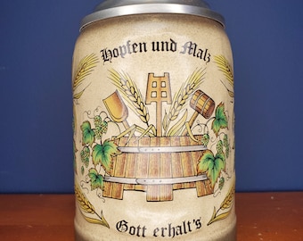May include: A beige ceramic beer stein with a silver lid. The stein features a brown wooden barrel with a scoop, a mallet, and wheat stalks. The text "Hopfen und Malz Gott erhalt's" is printed on the stein.