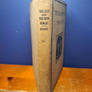 Może przedstawiać: Książka w kolorze beżowym z tytułem "The Old and The New Magic" autorstwa Evansa. Książka stoi pionowo na drewnianej powierzchni.