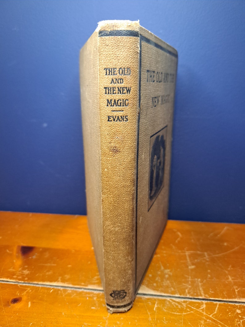 Może przedstawiać: Książka w kolorze beżowym z tytułem "The Old and The New Magic" autorstwa Evansa. Książka stoi pionowo na drewnianej powierzchni.