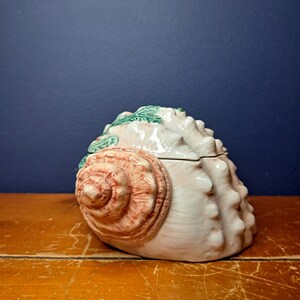 May include: A ceramic seashell trinket box with a pink and white shell design and green seaweed accents.
