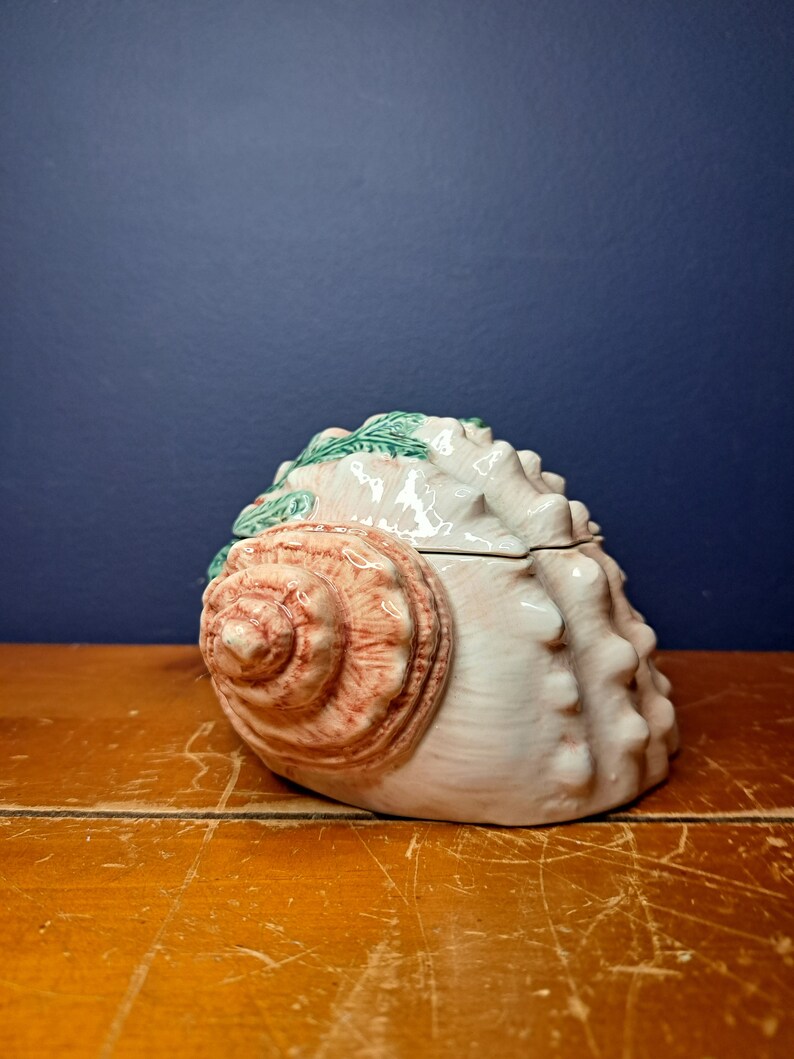 May include: A ceramic seashell trinket box with a pink and white shell design and green seaweed accents.