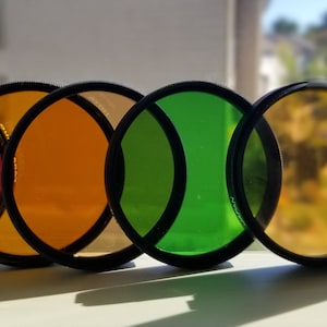 May include: A set of six round camera lens filters in red, orange, yellow, green, and two shades of yellow. The filters are arranged in a row, with the red filter on the left and the yellow filter on the right.