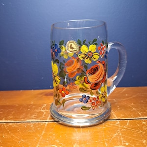 Könnte beinhalten: Ein klares Glasbierkrug mit einem Blumendesign. Das Design zeigt rote, orange, gelbe, blaue und grüne Blumen. Der Krug hat einen Henkel und einen runden Boden.