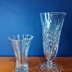 Puede incluir: Dos jarrones de cristal transparente. El jarr&oacute;n m&aacute;s peque&ntilde;o tiene una parte superior acampanada y un dise&ntilde;o acanalado vertical. El jarr&oacute;n m&aacute;s grande tiene un patr&oacute;n de corte de diamante y una base ancha y circular. Ambos jarrones est&aacute;n sobre una superficie de madera sobre un fondo azul.