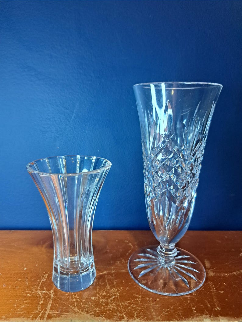 Puede incluir: Dos jarrones de cristal transparente. El jarr&oacute;n m&aacute;s peque&ntilde;o tiene una parte superior acampanada y un dise&ntilde;o acanalado vertical. El jarr&oacute;n m&aacute;s grande tiene un patr&oacute;n de corte de diamante y una base ancha y circular. Ambos jarrones est&aacute;n sobre una superficie de madera sobre un fondo azul.