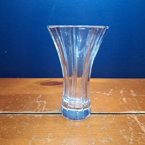 Puede incluir: Un jarr&oacute;n de cristal transparente con la parte superior acampanada y un dise&ntilde;o acanalado vertical. El jarr&oacute;n est&aacute; sobre una superficie de madera contra un fondo azul. El jarr&oacute;n est&aacute; vac&iacute;o y parece ser de cristal o vidrio de alta calidad.