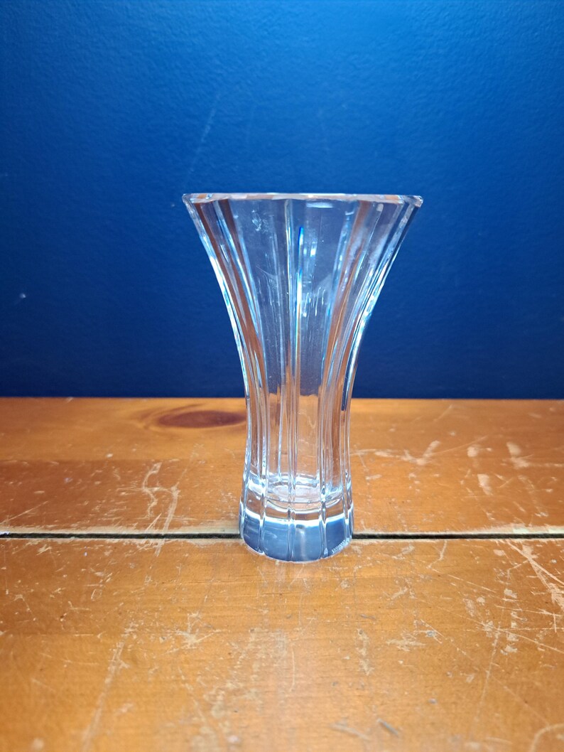 Puede incluir: Un jarr&oacute;n de cristal transparente con la parte superior acampanada y un dise&ntilde;o acanalado vertical. El jarr&oacute;n est&aacute; sobre una superficie de madera contra un fondo azul. El jarr&oacute;n est&aacute; vac&iacute;o y parece ser de cristal o vidrio de alta calidad.
