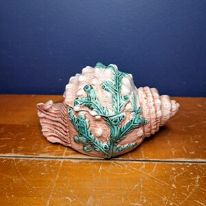 May include: A ceramic seashell trinket box with a pink and green glaze. The seashell is decorated with green seaweed and has a hinged lid.