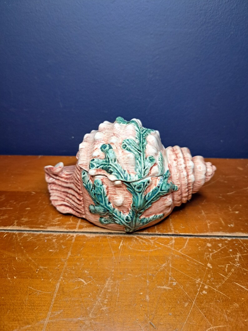 May include: A ceramic seashell trinket box with a pink and green glaze. The seashell is decorated with green seaweed and has a hinged lid.