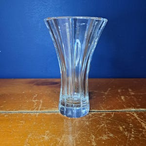 Puede incluir: Un jarr&oacute;n de cristal transparente con la parte superior acampanada y un dise&ntilde;o acanalado vertical. El jarr&oacute;n est&aacute; sobre un fondo azul y sobre una superficie de madera. El jarr&oacute;n mide unos 15 cm de alto.