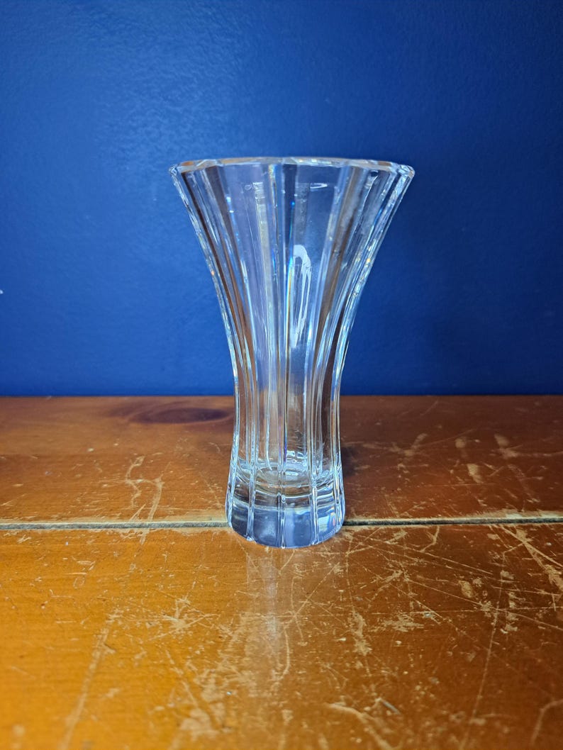 Puede incluir: Un jarr&oacute;n de cristal transparente con la parte superior acampanada y un dise&ntilde;o acanalado vertical. El jarr&oacute;n est&aacute; sobre un fondo azul y sobre una superficie de madera. El jarr&oacute;n mide unos 15 cm de alto.