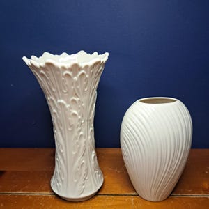 May include: Two white ceramic vases. One is tall with a ruffled top and a detailed, embossed leaf design. The other is a smaller, oval-shaped vase with a textured, vertical swirl pattern. Both vases are on a wooden surface against a dark blue background.