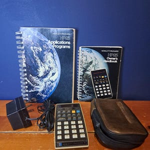 May include: A vintage Hewlett-Packard HP-25 calculator with its original accessories. The image includes the calculator, a power adapter, a carrying case, and two manuals titled "Applications Programs" and "Owner's Handbook". The calculator has a black and beige color scheme.