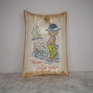 May include: A vintage ceramic dish with a cartoon illustration of a child fishing in a pond. The dish is decorated with a yellow flower, green reeds, and blue water. The text "Never drink water..." is printed on the dish.