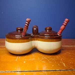 May include: A double ceramic condiment dish with brown and white glaze and two wooden spoons. The dish has a handle in the center.