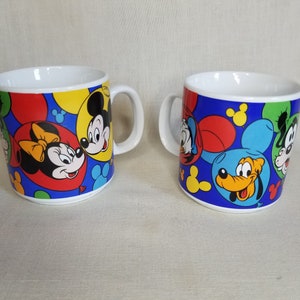 以下が含まれることがあります： 青い背景にカラフルなミッキーマウス、ミニーマウス、ドナルドダック、グーフィー、プルートの漫画の画像が描かれた白いセラミック製のマグカップ2つ。マグカップには持ち手があり、風船で飾られています。