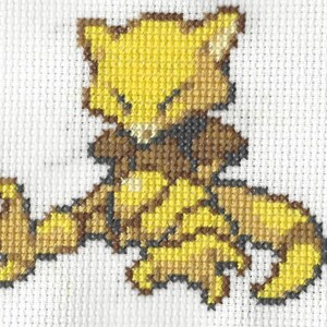 Könnte beinhalten: Kreuzstich von Abra, einem gelb-braunen Charakter aus der Pokémon-Serie. Der Kreuzstich befindet sich auf einem weißen Stoffhintergrund. Die Abra-Figur sitzt mit ausgestreckten Armen.