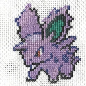 Puede incluir: Punto de cruz morado y verde de un personaje de Pokemon. El personaje tiene una oreja grande y puntiaguda y un ojo pequeño y redondo.