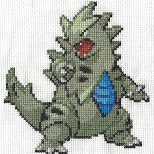 Può includere: Schema a punto croce di un personaggio Pokemon verde e blu. Il personaggio ha un occhio rosso e una schiena spinosa.