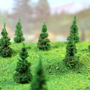 May include: A collection of miniature green pine trees on a green grassy surface. The trees are different sizes and are arranged in a random pattern.