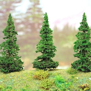 May include: Three miniature green model pine trees with detailed foliage stand on a grassy surface. The trees have a realistic appearance, with textured branches and a natural color palette, perfect for model train layouts or dioramas.