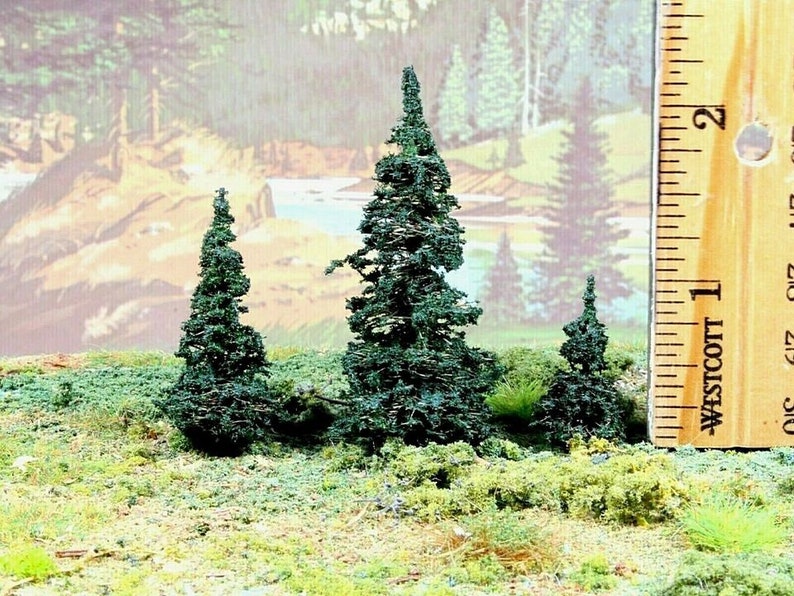 May include: Three miniature evergreen trees, approximately 2 inches tall, are arranged on a green, grassy surface. A wooden ruler with the text "Westcott" is visible in the background.