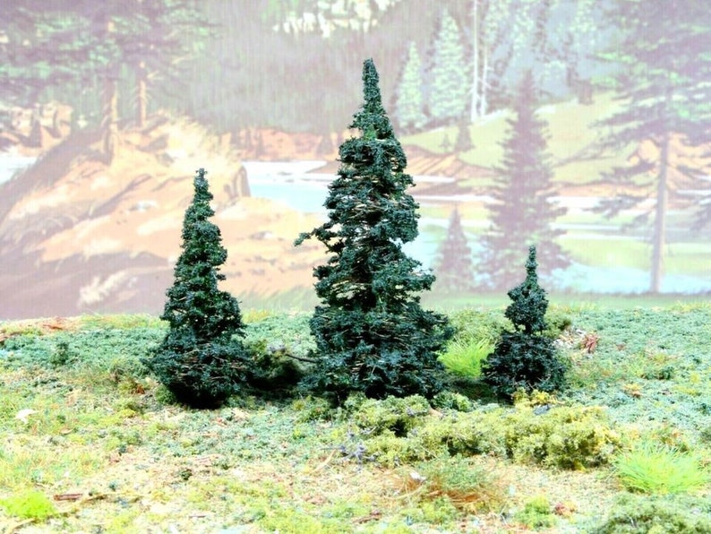 May include: Three miniature evergreen trees with dark green foliage, set against a painted backdrop of a forest scene.