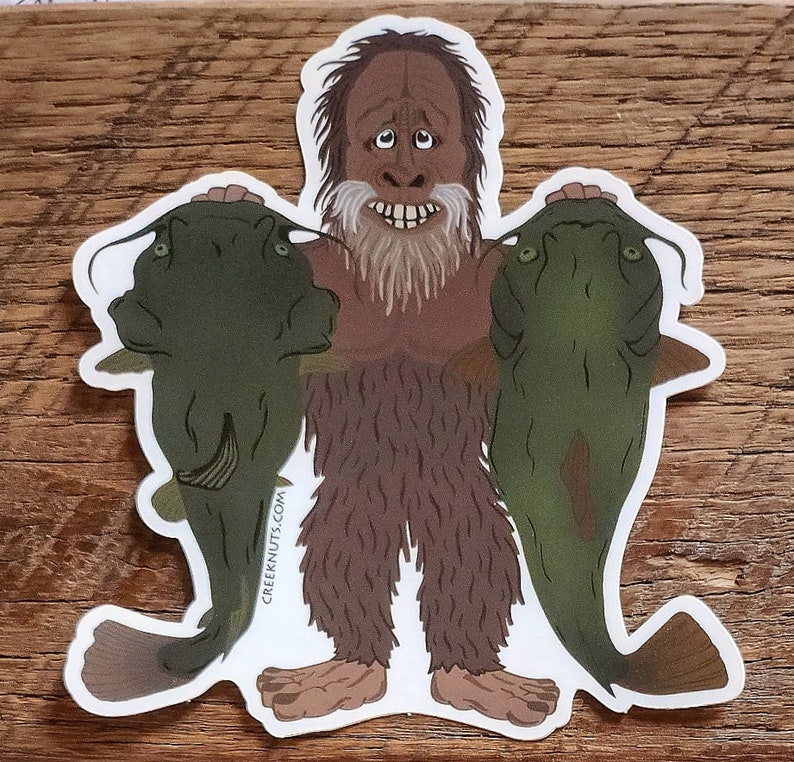 May include: A sticker featuring a cartoon illustration of a brown, hairy creature holding two green catfish. The creature has a long beard and a surprised expression. The sticker has the text "creeknuts.com" on the bottom left corner.