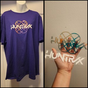 Peut inclure: Un t-shirt violet avec le mot "HUNTRX" en blanc, avec un motif or et turquoise au-dessus du texte. Le t-shirt est présenté sur un mannequin. Une main tient une feuille transparente avec le même motif.