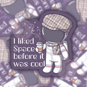 May include: A cartoon astronaut wearing a white spacesuit, a plaid hat, and a scarf, holding a cup of coffee. The astronaut is standing on a purple background with the text "I liked Space before it was cool".