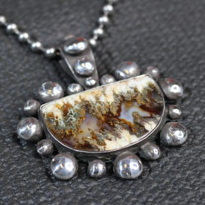 May include: A semi-circular pendant featuring a dendritic agate stone with brown, white, and translucent patterns, set in a textured silver frame with small spherical silver accents along the edge, suspended from a silver ball chain necklace.
