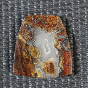 Könnte beinhalten: Ein polierter, rechteckiger Edelstein mit einer zentralen weißen Höhle. Der Stein weist eine Mischung aus Farben auf, darunter Rot, Braun, Gelb und Grau, wodurch ein natürliches, erdiges Muster entsteht. Der Edelstein liegt vor einem strukturierten grauen Hintergrund.