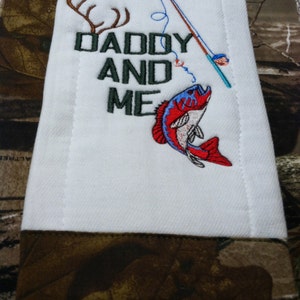 Op de afbeelding: Een witte spuugdoek met een bruine rand. Het heeft een geborduurd ontwerp van een hengel, een vis, geweien en de tekst "Daddy and Me".