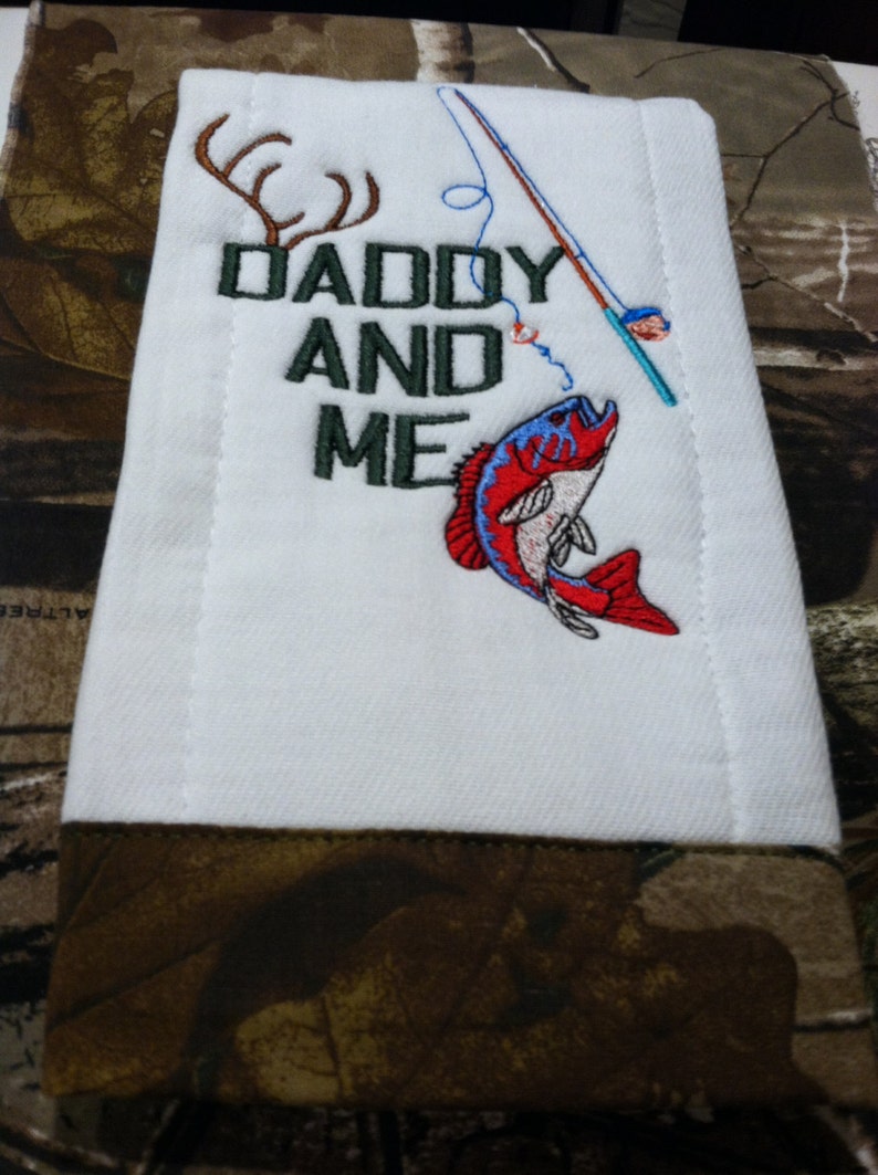 Op de afbeelding: Een witte spuugdoek met een bruine rand. Het heeft een geborduurd ontwerp van een hengel, een vis, geweien en de tekst "Daddy and Me".