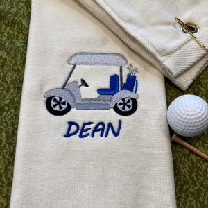 May include: A cream-colored golf towel featuring an embroidered light blue and grey golf cart with a blue seat and a blue golf bag containing clubs. The name DEAN is embroidered in blue thread below the cart. A white golf ball sits nearby.