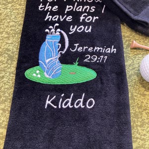 May include: A black golf towel featuring white embroidered text that reads For I know the plans I have for you Jeremiah 29:11 and Kiddo. The design includes a blue golf bag on a green patch of grass with white golf balls and a golf tee.