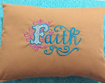 Op de afbeelding: Een bruin kussen met het woord "Faith" geborduurd in blauwe en roze cursieve letters met decoratieve krulletjes.