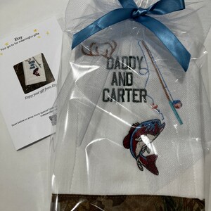 Op de afbeelding: Witte geborduurde keukendoek met een bruine camouflage rand. De handdoek heeft een hengel, een vis en de tekst "Daddy and Carter".