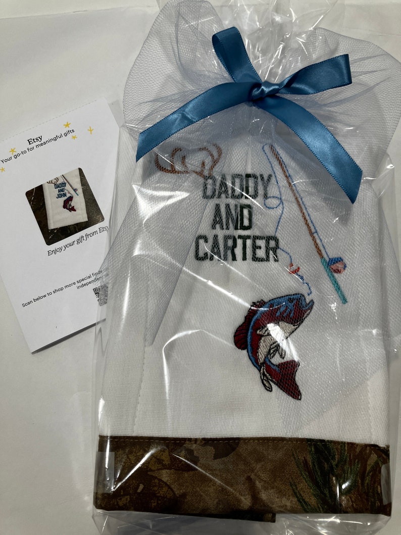 Op de afbeelding: Witte geborduurde keukendoek met een bruine camouflage rand. De handdoek heeft een hengel, een vis en de tekst "Daddy and Carter".