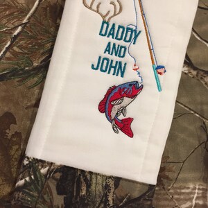 Op de afbeelding: Een witte spuugdoek met geborduurde tekst "Daddy and John" en een hengel met een rode en blauwe vis. De achtergrond is een camouflagepatroon.