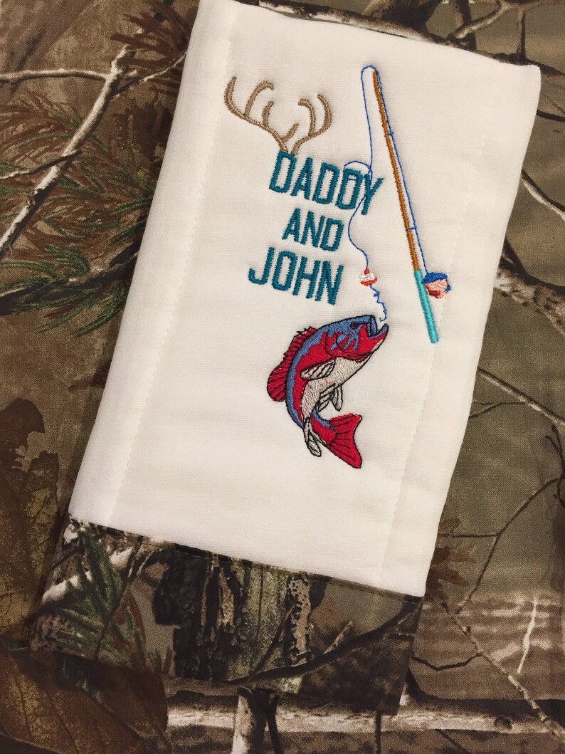 Op de afbeelding: Een witte spuugdoek met geborduurde tekst "Daddy and John" en een hengel met een rode en blauwe vis. De achtergrond is een camouflagepatroon.