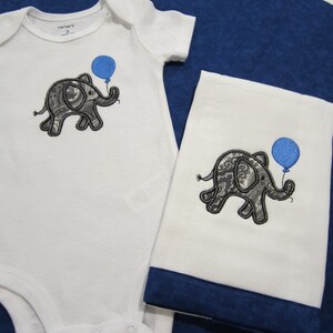 May include: A white onesie with a gray elephant holding a blue balloon embroidered on the front. A white burp cloth with a gray elephant holding a blue balloon embroidered on the front. The burp cloth has a blue trim.