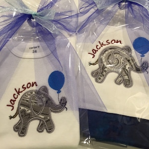 May include: Two white baby onesies with blue and purple tulle bows. The onesies feature an embroidered elephant with a blue balloon and the name "Jackson" in red lettering. One onesie has a "carter's" tag with the size "24" printed on it.