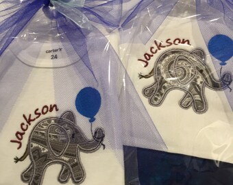 Op de afbeelding: Twee witte baby-rompers met blauwe en paarse tule strikken. De rompers hebben een geborduurde olifant met een blauwe ballon en de naam "Jackson" in rode letters. Eén romper heeft een "carter's" label met de maat "24" erop.