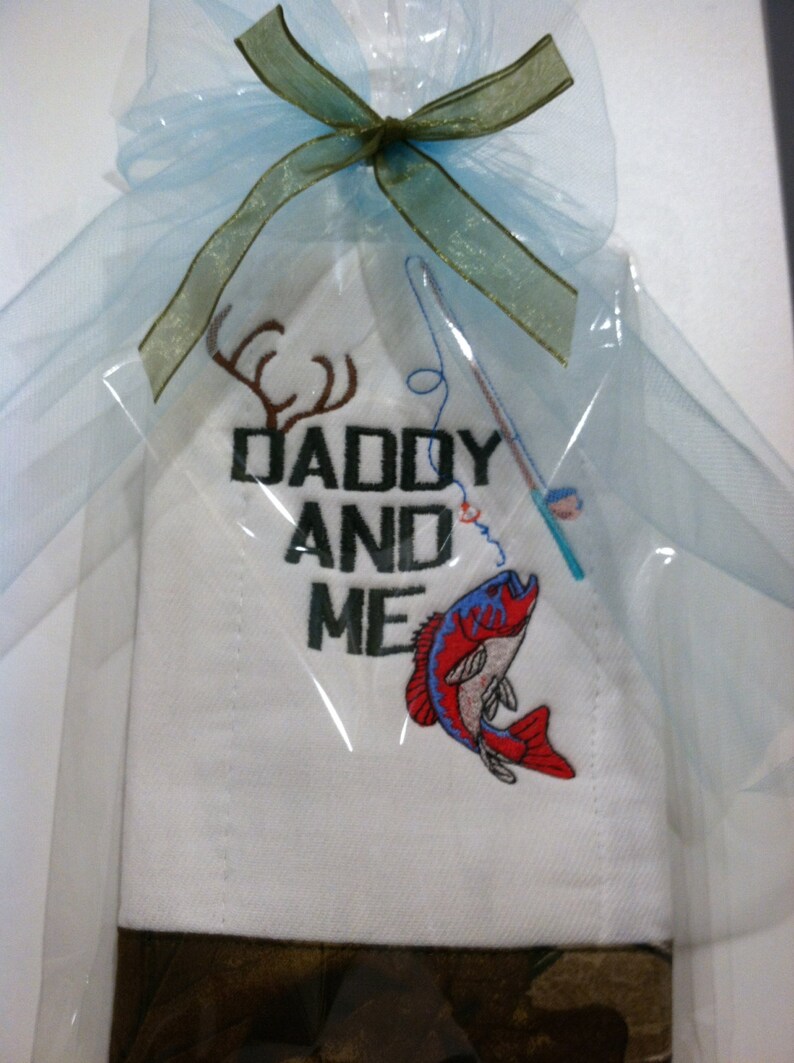 Op de afbeelding: Een witte geborduurde keukendoek met de tekst "Daddy and Me" en een rode vis met een hengel en een hertengewei. De handdoek is verpakt in een blauw en groen doorschijnend lint en verpakt in een doorzichtige plastic zak.
