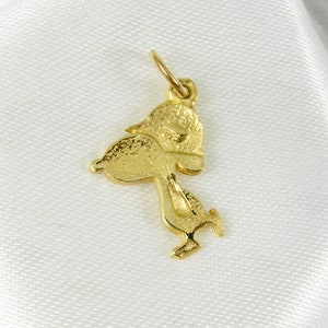 May include: A gold-colored charm in the shape of a cartoon character, possibly a duck or bird, with a textured surface. The charm has a loop at the top for attachment to a necklace or bracelet. The charm is set against a white background.
