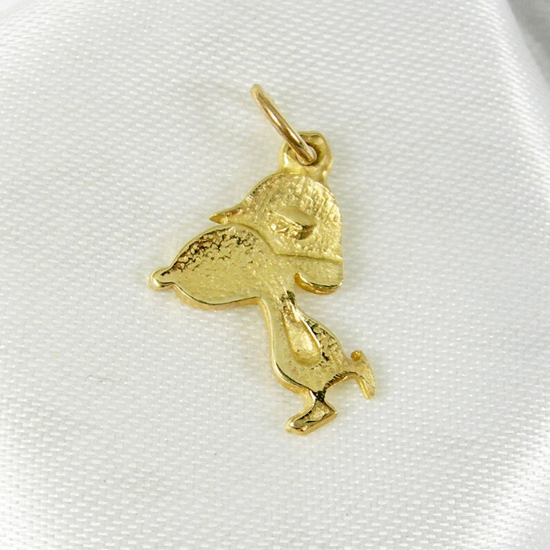 May include: A gold-colored charm in the shape of a cartoon character, possibly a duck or bird, with a textured surface. The charm has a loop at the top for attachment to a necklace or bracelet. The charm is set against a white background.
